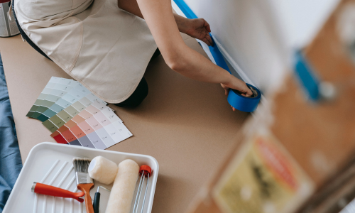 A woman laying down paint tape at the bottom of a wall preparing to paint with samples and supplies next to her
